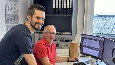 Foto von zwei Männern am PC-Arbeitsplatz. Beide lächeln in die Kamera.
