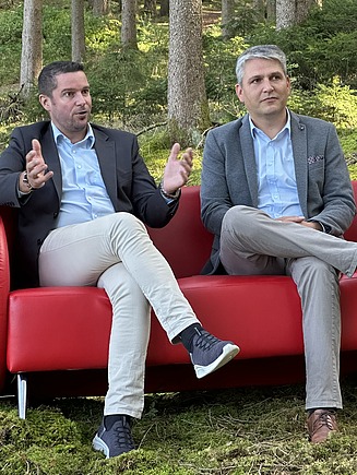 Foto von zwei Männern sitzend auf einer roten Couch inmitten des Waldes