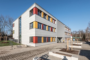 Außenaufnahme der Schule in Erfurt. Man sieht ein großes, weißes Gebäude mit bunten Farbelementen bei den Fenstern.