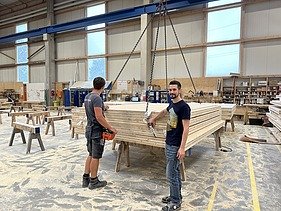Foto von zwei Männern in der Produktionshalle. Sie stehen vor einem Stapel voller Holzelemente, welche am Kran hängen.