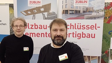 Foto von zwei Männern. Im Hintergrund sind Ausschnitte zweier Roll-Ups zu sehen. 