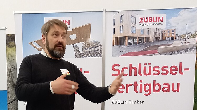 Acquisition at ZÜBLIN Timber Photo of a man in dark clothing gesticulating with his hands in front of two roll-ups.
