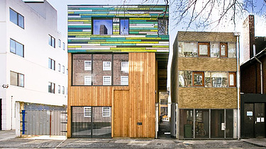 Foto mehrerer Gebäude. In der Mitte ist ein neuer Holzbau mit bunter Fassade im oberen Teil zu sehen.