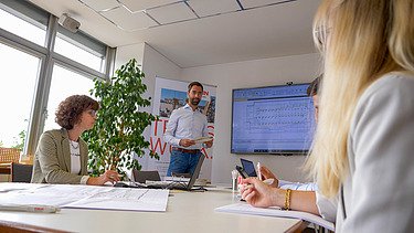Photo of a meeting room with three seated and one standing person.