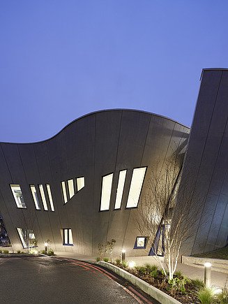 Außenansicht des Maggie Center in London. Man sieht ein geschwungenes Gebäude aus Holz in der Nacht.