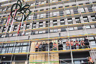 News ZÜBLIN Timber. Topping-out ceremony for the T3 office in Aichach. Fabian Schön, carpenter, found the right words for the timber construction work. Photo of the topping-out ceremony for the new T3 office building in Aichach. The topping-out wreath is raised. People are standing on the scaffolding.