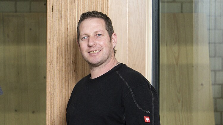 Photo of a young man standing in front of a wooden element with glass panes.