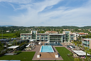 Photo of the Hotel Aqualux ini Bardolino. You can see a large building with a glass façade and flat roof, as well as a pool.