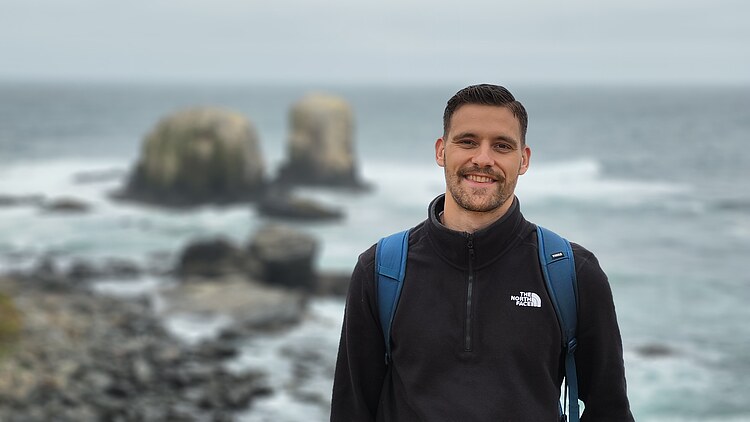 Photo of a man in black clothing. The sea and rocks can be seen in the background.