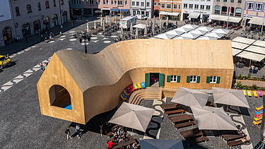 Foto des NEXT500-Pavillons auf dem Augsburger Marktplatz. Sie können ein modernes Holzgebäude sehen.