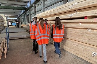 Photo of several young women walking past a stack of LENO timber in the ZÜBLIN Timber production hall.