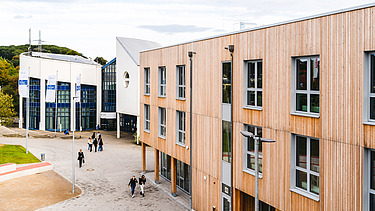Foto der Uni Witten von außen. Man sieht ein großes Gebäudes aus Holz.
