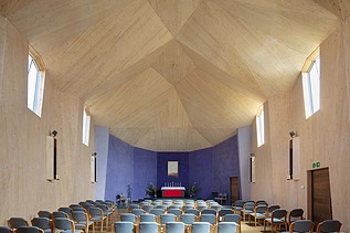 Foto eines Innenraums einer Kapelle mit großer gewölbter Holzdecke. Eine Seite hat eine blaue Wand.