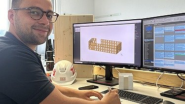 Foto von einem jungen Mann, der an einem Bildschirmarbeitsplatz sitzt und in die Kamera lächelt.