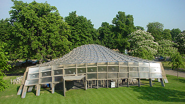 Foto des Serpentine Pavillons in London. Man sieht eine offene, begehbare Holzkonstruktion auf einer grünen Wiese mit einem Baum.
