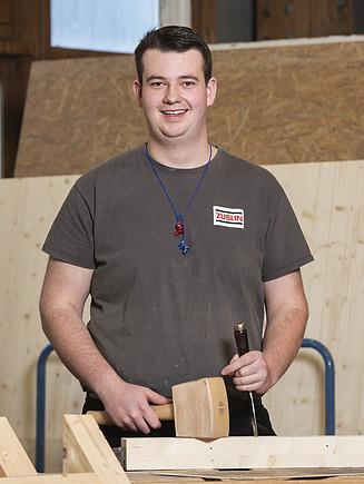 Foto von einem jungen Zimmerer der an einem kleinen Holzelement arbeitet.