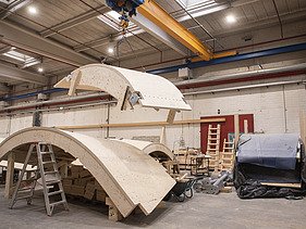 Photo of a production hall with two curved wooden shells, one hanging from the crane