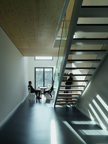Foto eines Innenraumes mit Treppenaufgang. Menschen sitzen und stehen am Fenster.