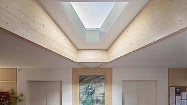 Foto eines Innenraumes mit Holzdecke und dreieckigem Fenster. Ein Bild hängt an der Wand.