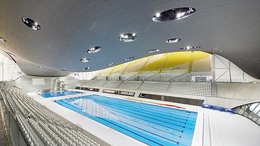 Photo of the Aquatics Center in London from the inside. You can see the wooden roof construction and a large swimming pool.