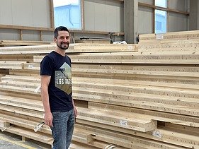 Foto eines jungen Mannes, der in der Produktion vor einem Holzplattenstapel steht und lächelt