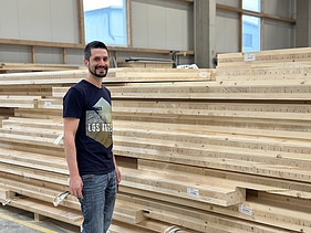 Photo of a young man standing in front of a stack of wooden panels in production and smiling