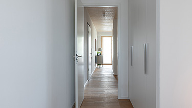 Foto eines leeren, weißen Flures im Holzhochhaus CARL. Man sieht Holzoberflächen auf dem Boden und an der Decke