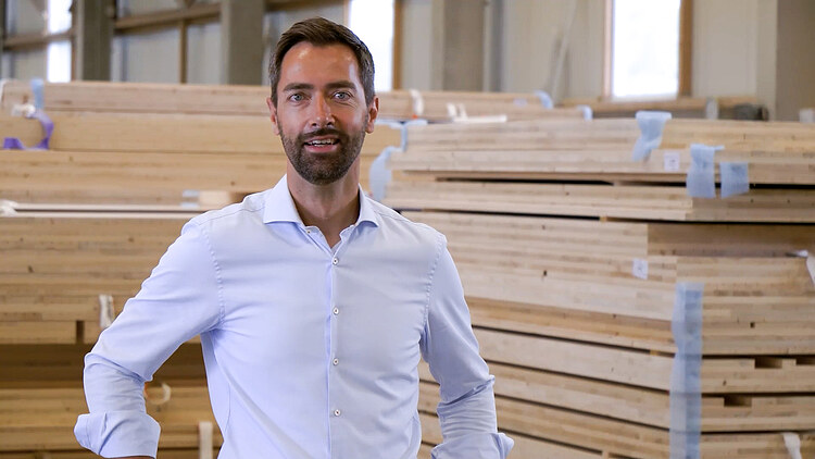 Photo of a man in a white shirt standing in front of a stack of LENO wood in production