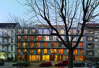 Foto eines mehrgeschossigen Gebäudes in dämmeriger Lichtstimmung. Zuvor stehen blattlose Bäume.