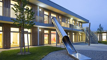 Außenaufnahme der Kindertagesstätte Campeon Infineon in Neubiberg. Man sieht ein Holzgebäude  und eine große Rutsche vom Haus in den Garten.