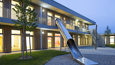 Exterior shot of the Campeon Infineon daycare center in Neubiberg. You can see a wooden building and a large slide from the house into the garden.