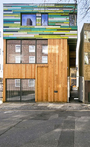 Foto mehrerer Gebäude. In der Mitte ist ein neuer Holzbau mit bunter Fassade im oberen Teil zu sehen.