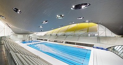 Photo of the Aquatics Center in London from the inside. You can see the wooden roof construction and a large swimming pool.