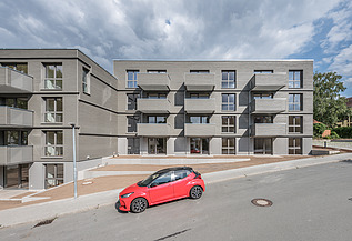 Foto der Joseph-Stiftung in Bayreuth. Man sieht ein Gebäude mit grauer Fassade und ein vorbeifahrendes, rotes Auto.