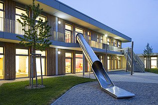 Außenaufnahme der Kindertagesstätte Campeon Infineon in Neubiberg. Man sieht ein Holzgebäude  und eine große Rutsche vom Haus in den Garten.