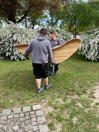 Foto von der Beförderung der Holzliegen von ZÜBLIN Timber. Zwei junge Männer tragen sie auf eine Wiese.