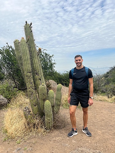 Photo of a young man in short black trousers and a black T-shirt and he is standing in the desert. He is standing next to a large cactus.