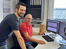 Photo of two men at a PC workstation. Both are smiling into the camera.