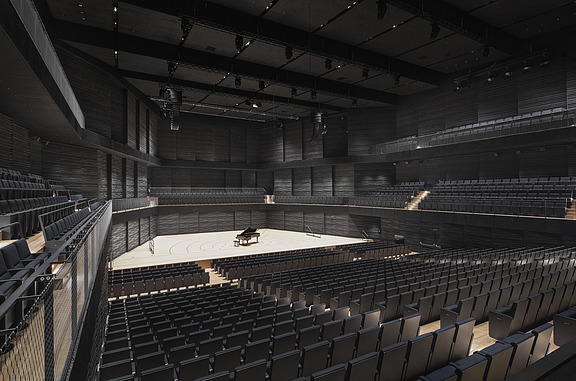 Photo of an interior of the interim venue Gasteig in Munich. You can see black wooden acoustic elements on the walls and ceiling and a grand piano in the middle of the stage.