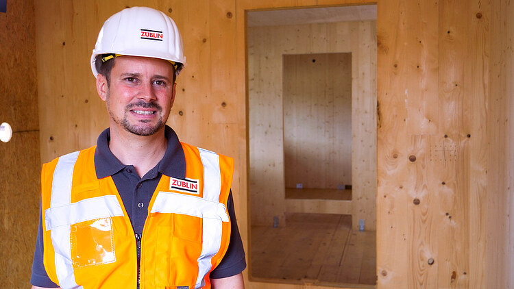 Picture of a young project manager in work clothes, smiling at the camera and standing in an interior made of LENO cross-laminated timber.