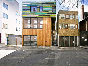 Foto mehrerer Gebäude. In der Mitte ist ein neuer Holzbau mit bunter Fassade im oberen Teil zu sehen.