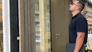 Foto von einem jungen Mann der vor einem Holzelement in der Produktion seht und es betrachtet.