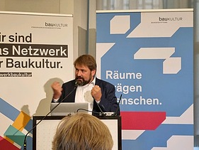 Photo of a man at the lectern. Roll-ups can be seen in the background.