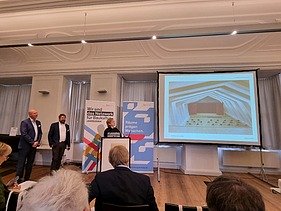 Foto von drei Personen, die ein Projekt präsentieren und vor Publikum stehen.