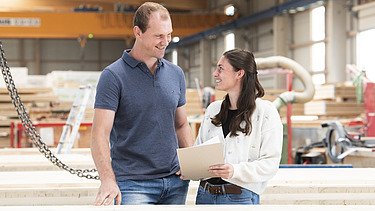 Foto eines großen Mannes und einer jungen Frau mit dunklen, langen Haaren, die in einer Produktionshalle stehen und sich anschauen.