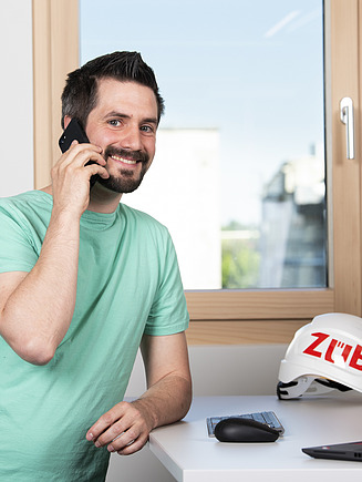 Photo of a young man standing at his PC workstation and talking on his cell phone.