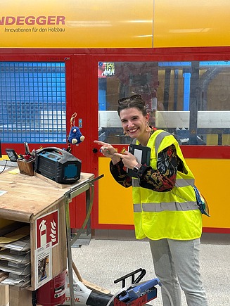 Eine Frau in der Fertigungshalle von ZÜBLIN Timber. Sie zeigt auf ein Maskottchen.