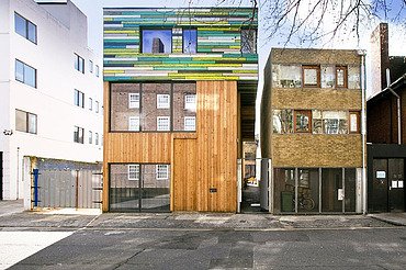 Foto mehrerer Gebäude. In der Mitte ist ein neuer Holzbau mit bunter Fassade im oberen Teil zu sehen.