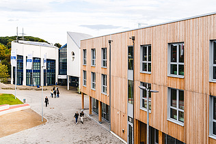 Foto der Uni Witten von außen. Man sieht ein großes Gebäudes aus Holz.