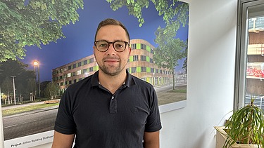 Foto von einem jungen Mann. Im Hintergrund sieht man ein Bild mit einem Holzgebäude darauf.